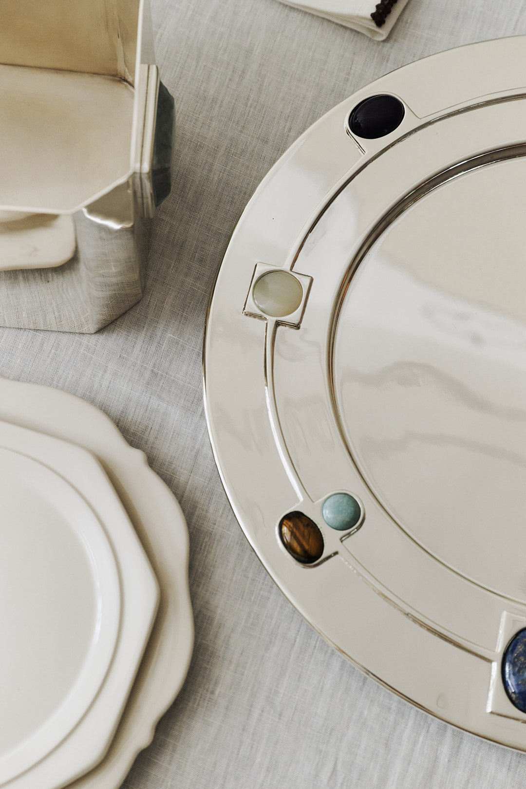 The Verola Round Tray, a silver amaro serving dish with colorful stone inlays, sits on a light tablecloth next to white ceramic plates and a metallic container.