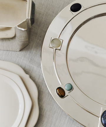 The Verola Round Tray, a silver amaro serving dish with colorful stone inlays, sits on a light tablecloth next to white ceramic plates and a metallic container.