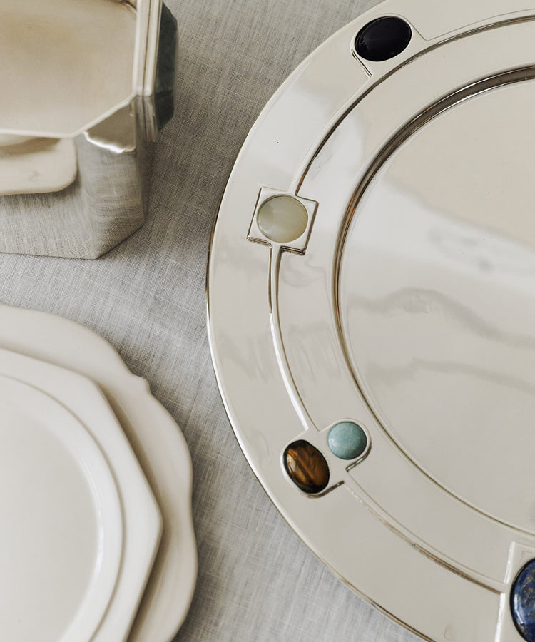 The Verola Round Tray, a silver amaro serving dish with colorful stone inlays, sits on a light tablecloth next to white ceramic plates and a metallic container.