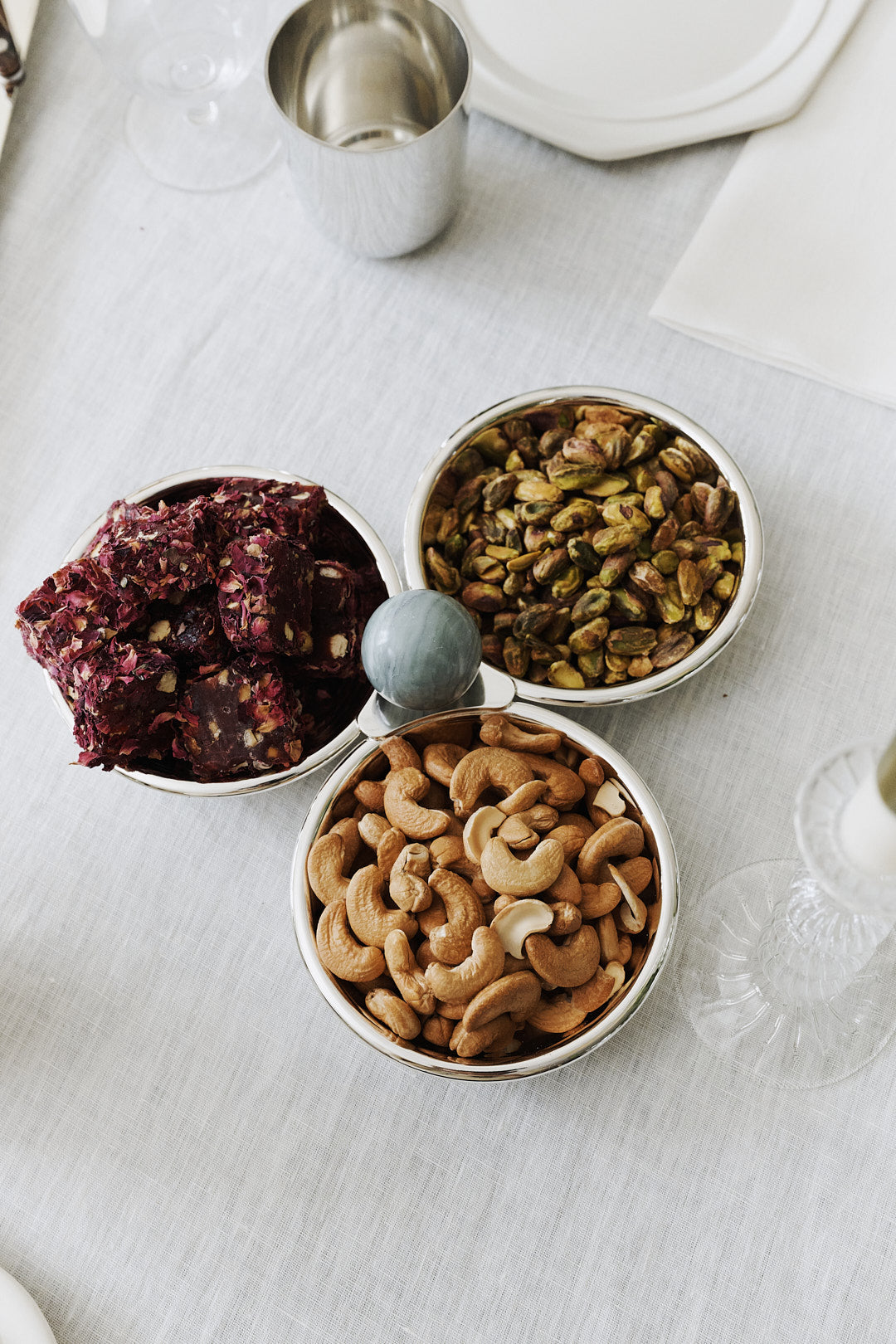 The Paros Triple Bowl Set features three silver bowls on a white tablecloth, filled with dried fruit squares, shelled pistachios, and cashew nuts—perfect as an elegant aperitivo serving set.