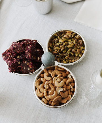 The Paros Triple Bowl Set features three silver bowls on a white tablecloth, filled with dried fruit squares, shelled pistachios, and cashew nuts—perfect as an elegant aperitivo serving set.