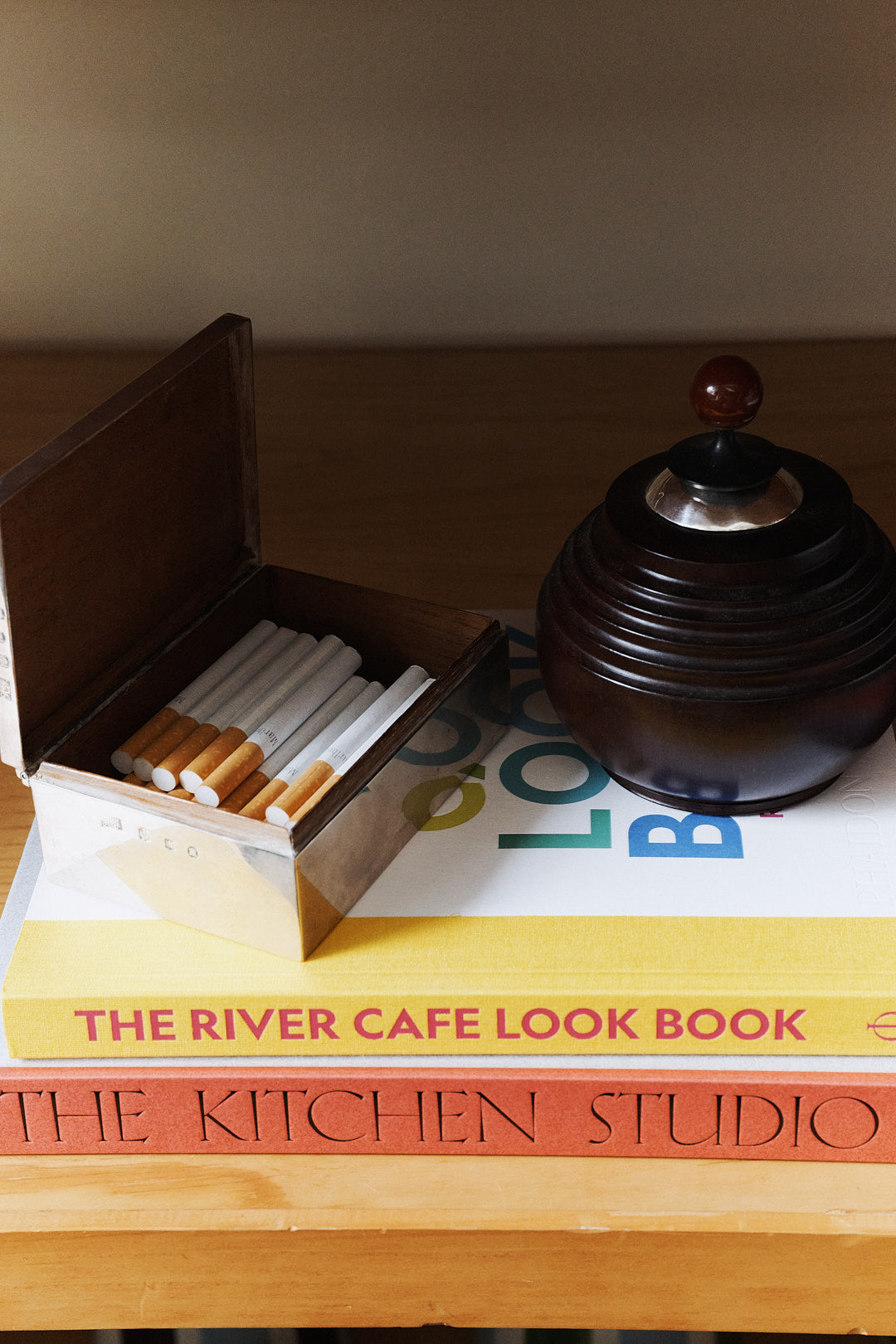 A metal cigarette box and a dark round ashtray rest atop two stacked books, titled The River Cafe Look Book and The Kitchen Studio: Culinary Creations by Artists—both celebrated art cookbooks that showcase contemporary artists and their unique recipes.