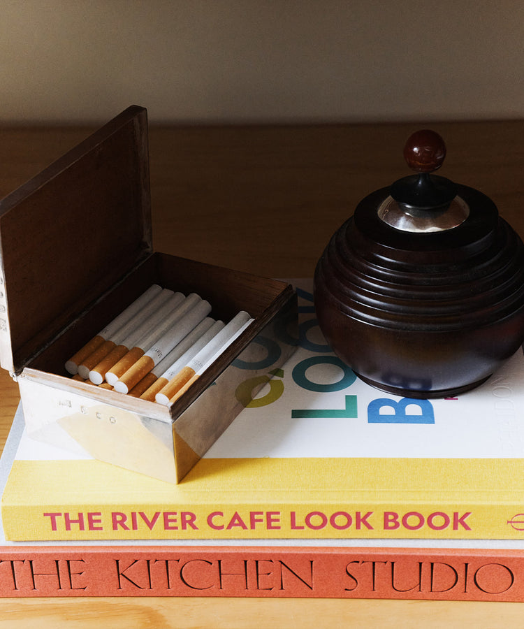 A metal cigarette box and a dark round ashtray rest atop two stacked books, titled The River Cafe Look Book and The Kitchen Studio: Culinary Creations by Artists—both celebrated art cookbooks that showcase contemporary artists and their unique recipes.