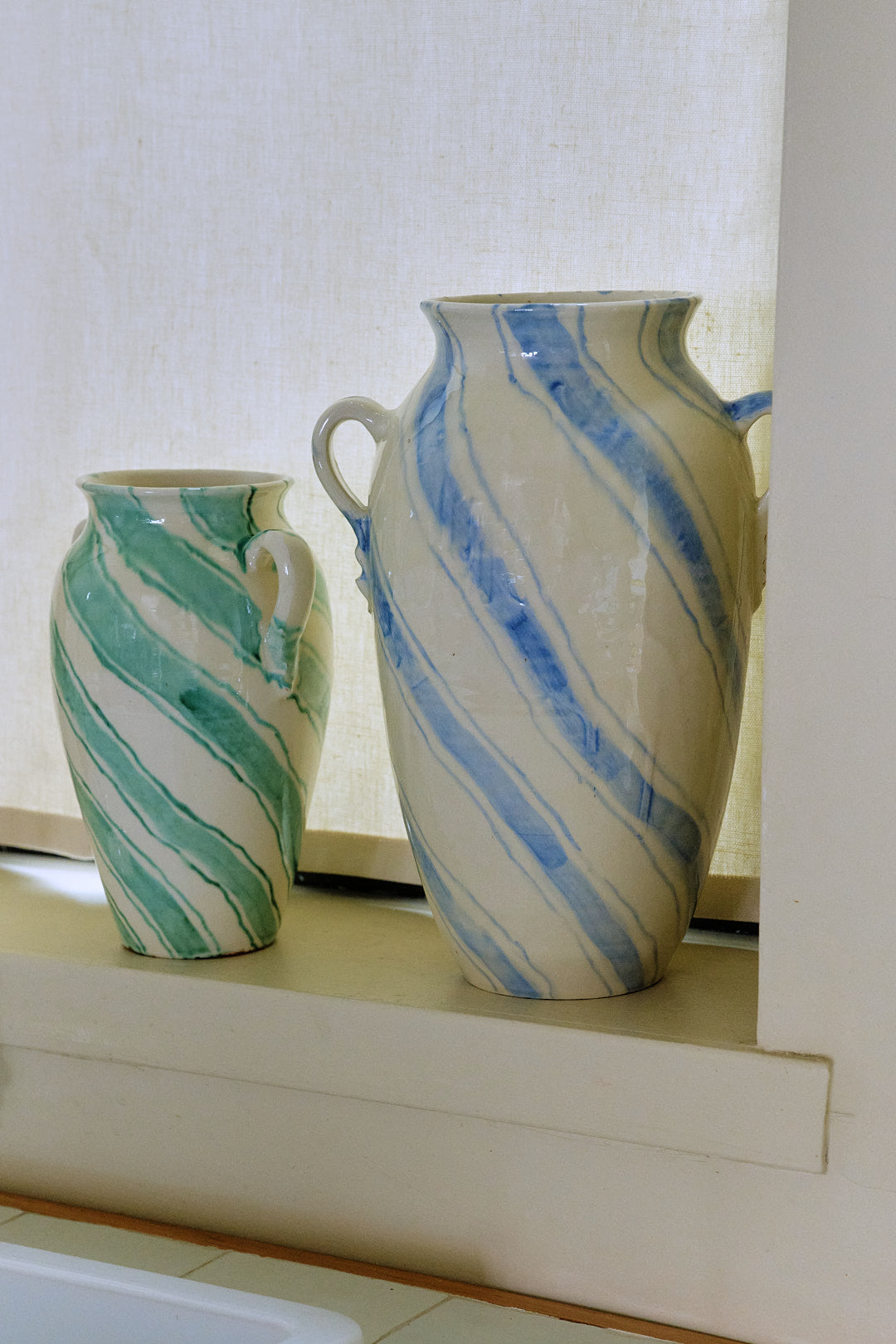 Two Ida Vases with diagonal stripes—one amphora-shaped in green and the other in blue—are displayed on a windowsill against a light curtain, adding a touch of modernist home decor to the space.