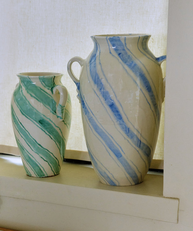 Two Ida Vases with diagonal stripes—one amphora-shaped in green and the other in blue—are displayed on a windowsill against a light curtain, adding a touch of modernist home decor to the space.