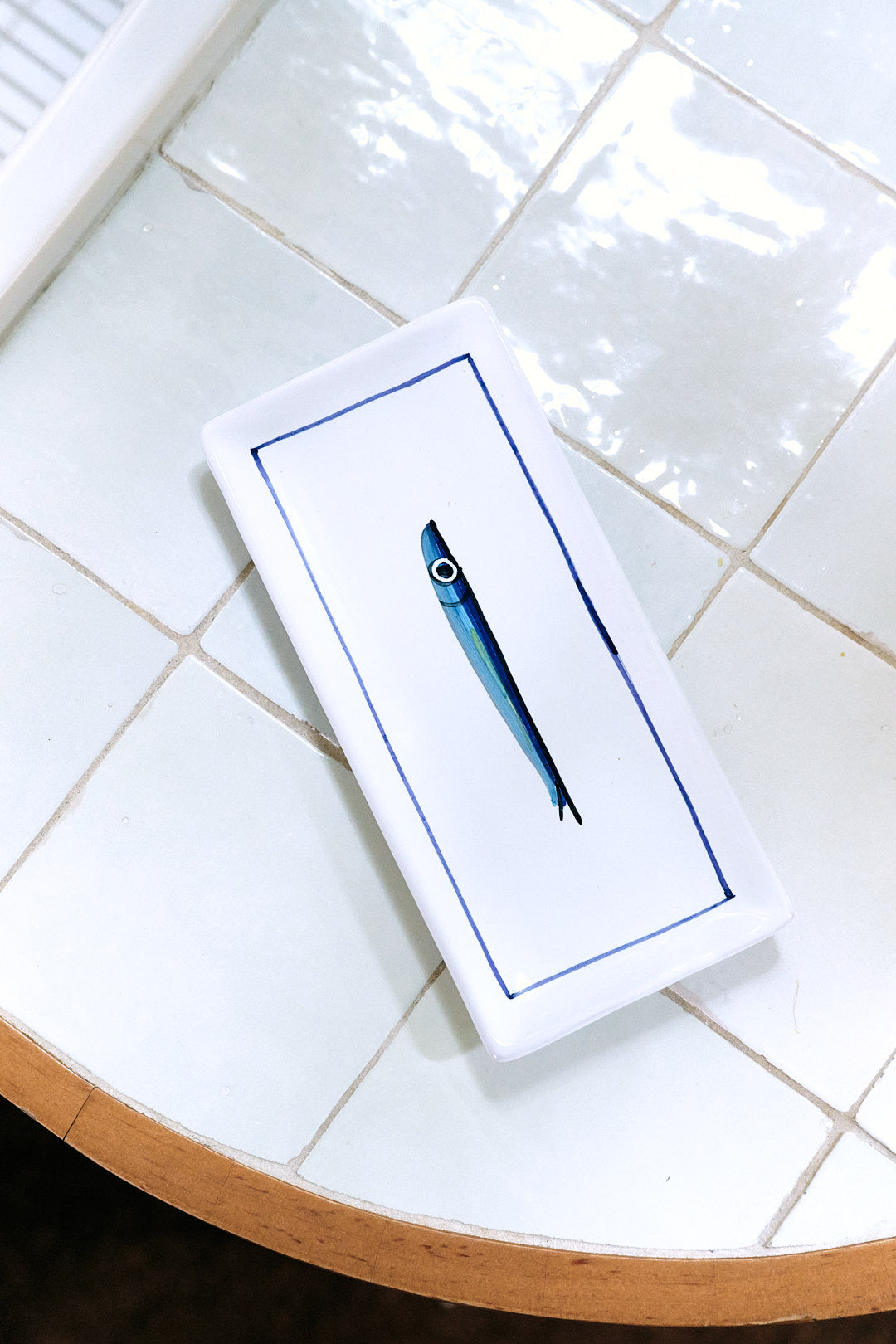 The Tommaso Rectangle Tray, a white ceramic serving tray with a hand-painted cobalt blue fish illustration in the center, is placed on a white tiled surface near the edge of a round table.