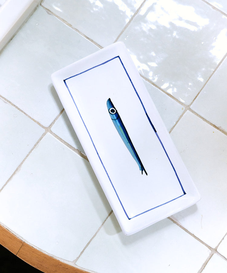 The Tommaso Rectangle Tray, a white ceramic serving tray with a hand-painted cobalt blue fish illustration in the center, is placed on a white tiled surface near the edge of a round table.