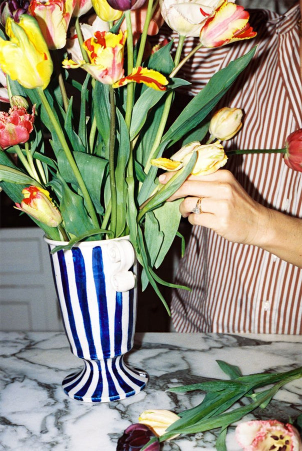 A person in a striped shirt arranges colorful tulips in a Thallo Pedestal Vase on a marble countertop.