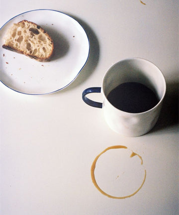 A Tea Mug filled with dark coffee sits beside a half-eaten slice of bread on a plate atop a table dotted with crumbs and coffee rings, showcasing minimalist design.