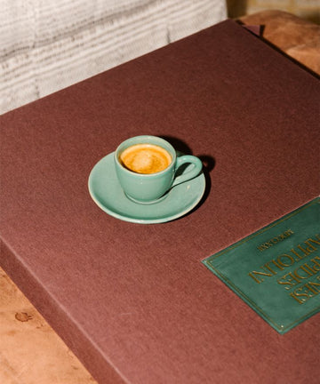 The Gio Espresso Set features a handmade ceramic mug, shown resting on a large brown book with a green label atop a wooden table.