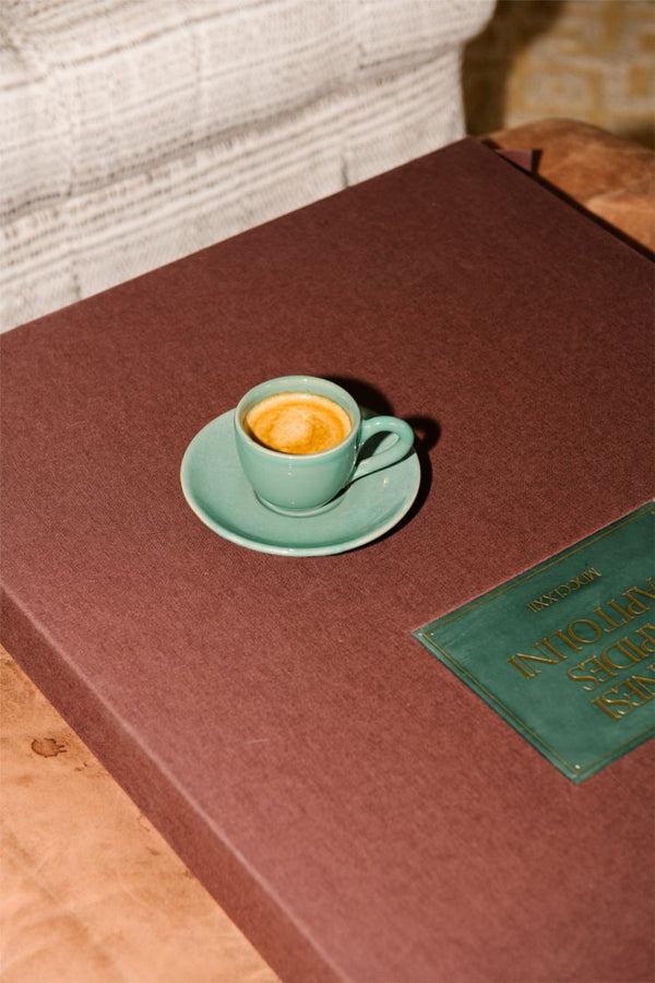 The Gio Espresso Set features a handmade ceramic mug, shown resting on a large brown book with a green label atop a wooden table.