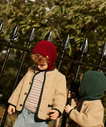 Two young children stand by a black metal fence outdoors, wearing Austrian Wool Jackets in beige with Nehru collars and cozy knit hoods—one red, one green—some crafted from soft merino wool. Trees form the background.