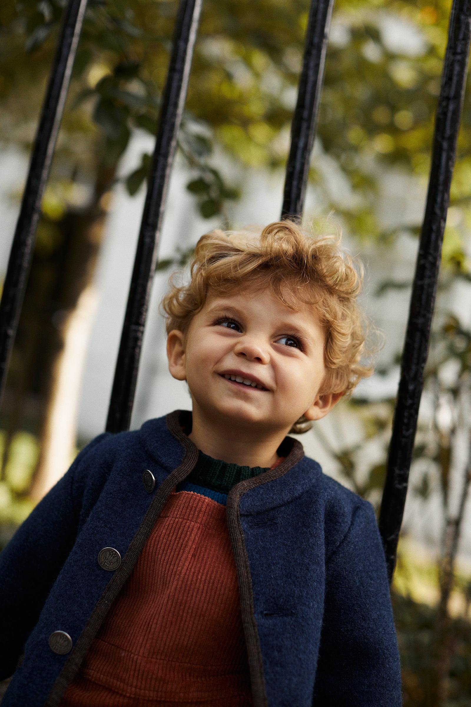 A young child with curly blonde hair, wearing a blue Austrian Wool Jacket and orange overalls, stands outdoors in front of a black metal fence and greenery.