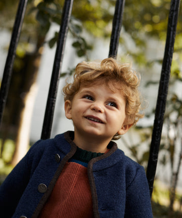 A young child with curly blonde hair, wearing a blue Austrian Wool Jacket and orange overalls, stands outdoors in front of a black metal fence and greenery.