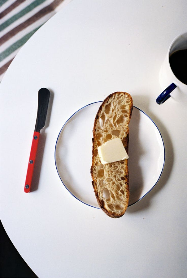 A slice of bread with butter rests on a plate beside a red-handled knife and black coffee, all atop a white table—perfectly complemented by tea served on our Cake Plate, Set of 4.