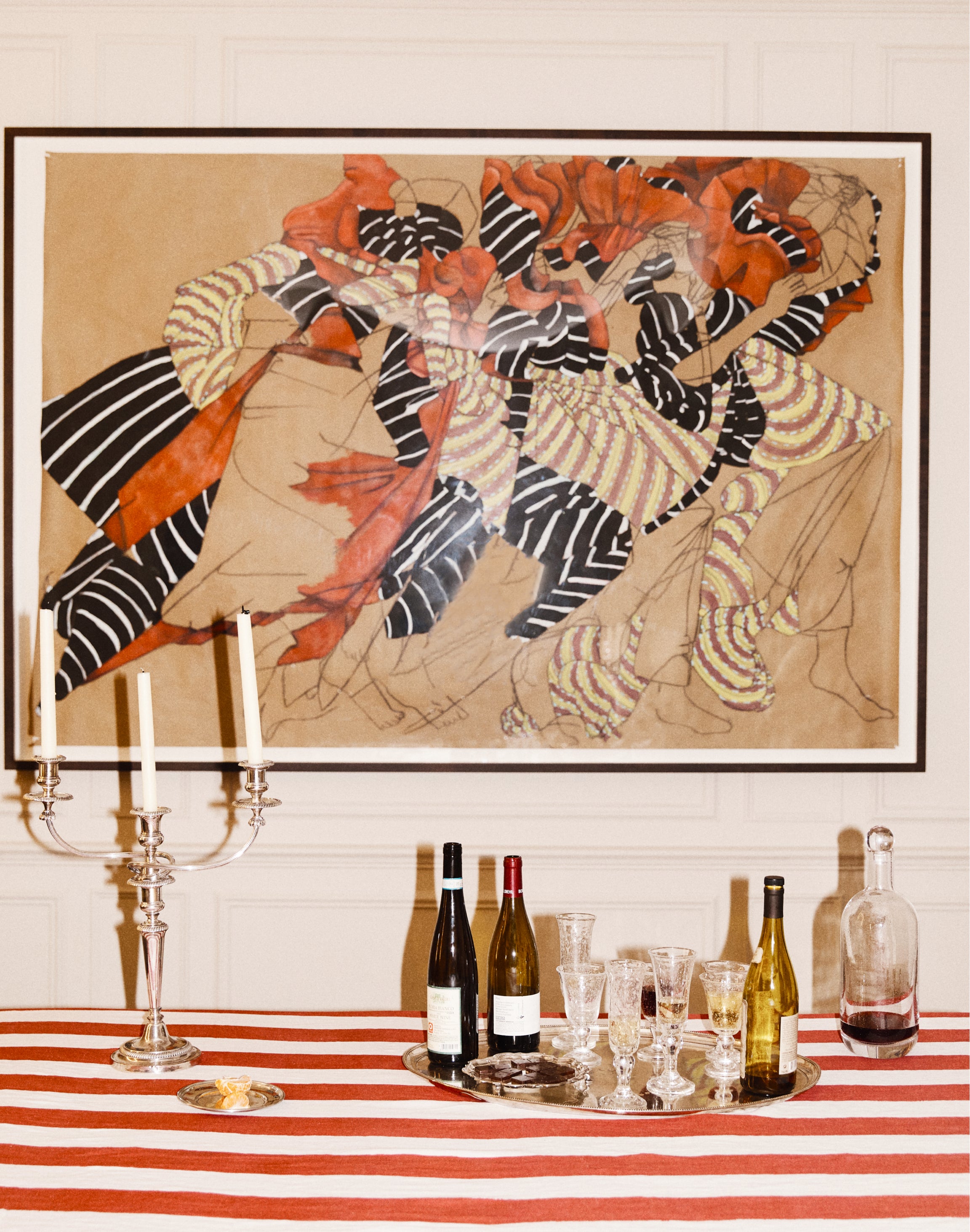 A Striped Tablecloth in festive red and white covers the table, which holds wine bottles, glasses, a decanter, and a silver candelabra; above hangs a large abstract painting.