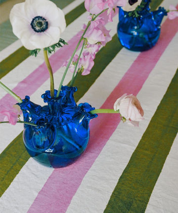 Two Lola Tulip Vases in blue glass, each filled with white and pink flower stems, sit on a green, pink, and white striped tablecloth—ideal for adding a charming touch to your home decor.
