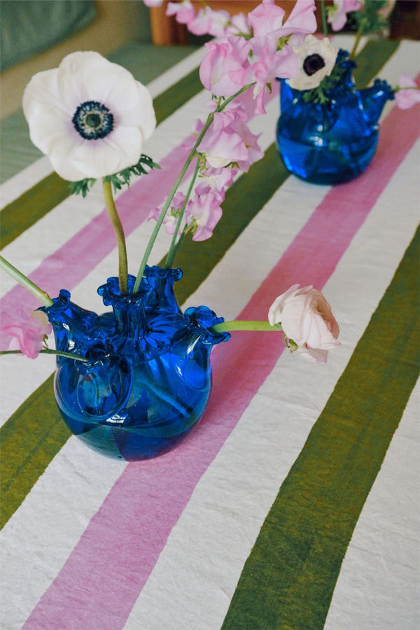 Two Lola Tulip Vases in blue glass, each filled with white and pink flower stems, sit on a green, pink, and white striped tablecloth—ideal for adding a charming touch to your home decor.
