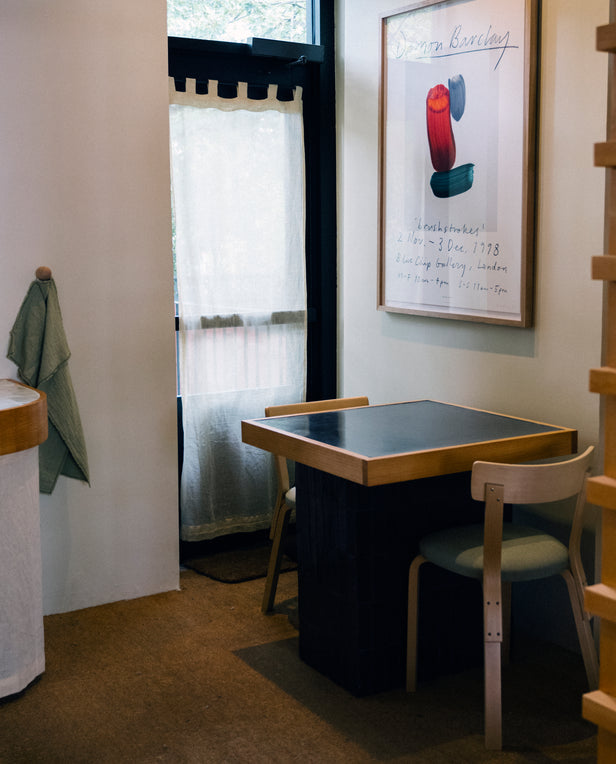 Small dining table with two chairs beside a wall, featuring a large framed art print and a window with sheer curtains.
