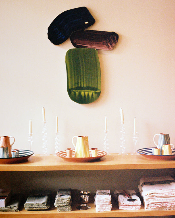 Wooden shelving unit with bowls, plates, and folded linens, set against a wall with three framed floral prints in red tones.