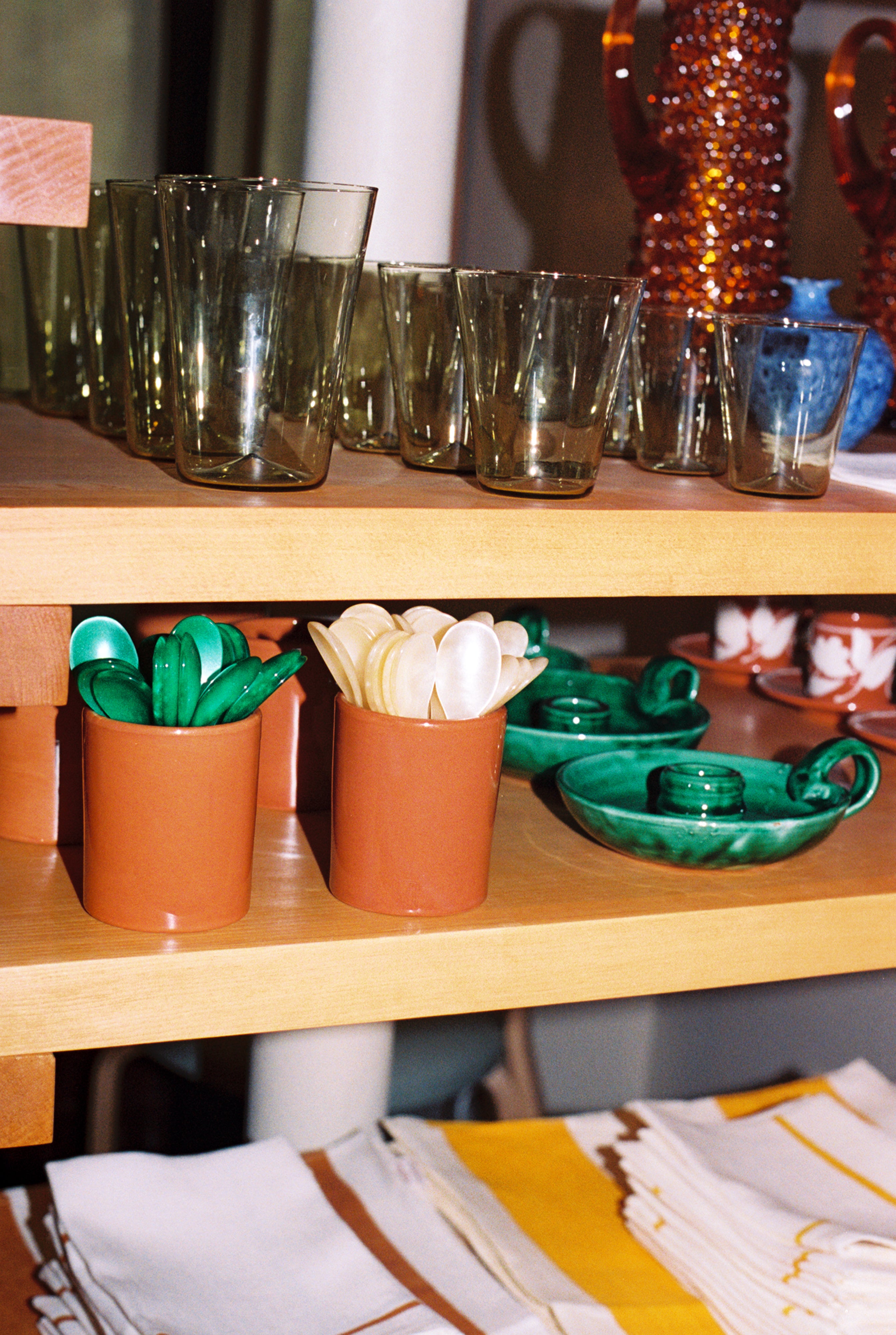 The Piccolo Spoon sits beside green ceramic plates and folded napkins, with drinking glasses and utensils in terracotta holders, all neatly arranged on wooden shelves.