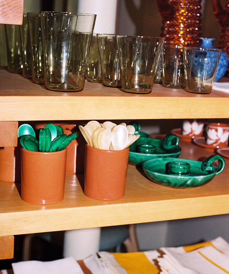 The Piccolo Spoon sits beside green ceramic plates and folded napkins, with drinking glasses and utensils in terracotta holders, all neatly arranged on wooden shelves.