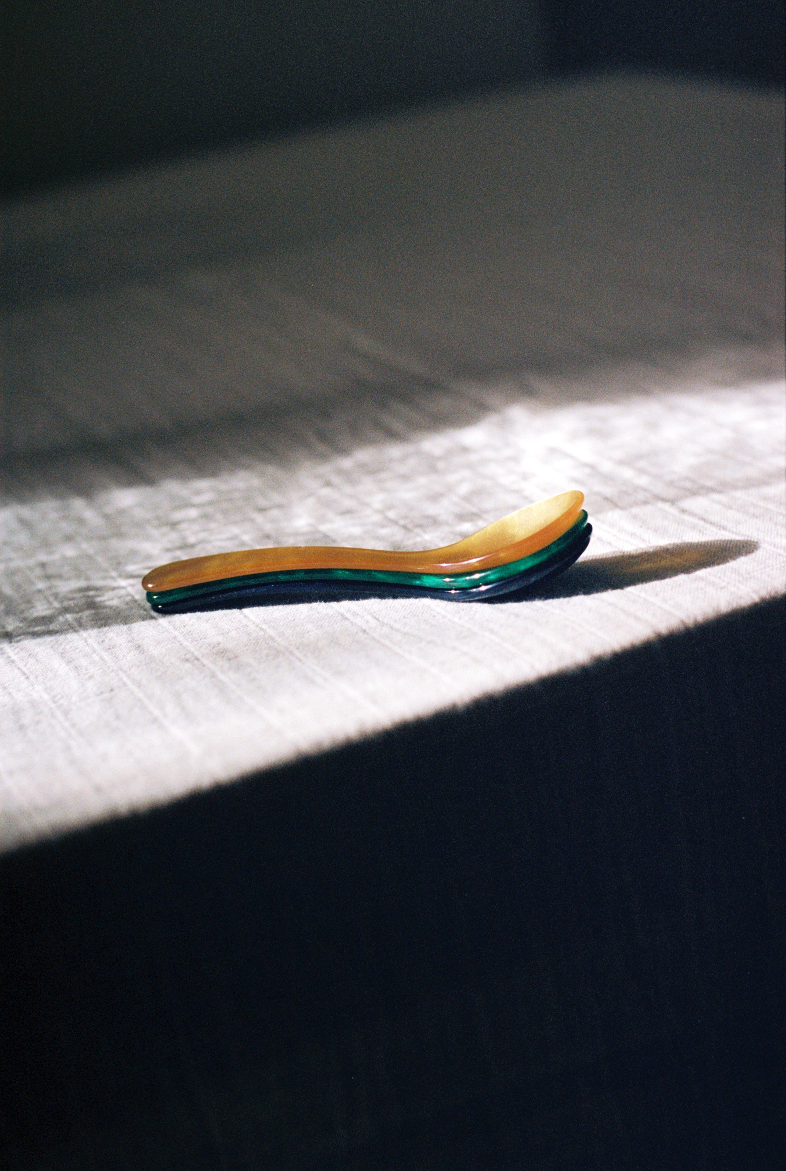 The Piccolo Spoon rests on a textured, sunlit tablecloth with its shadow to the right—ideal for soft boiled eggs and conveniently dishwasher safe.