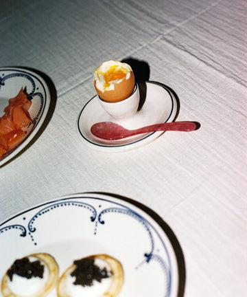 A soft-boiled egg in an egg cup with a pink Piccolo Spoon, beside plates of smoked salmon and blinis with cream and caviar on a white tablecloth. The dishwasher-safe Piccolo Spoon adds style and convenience to this elegant breakfast spread.