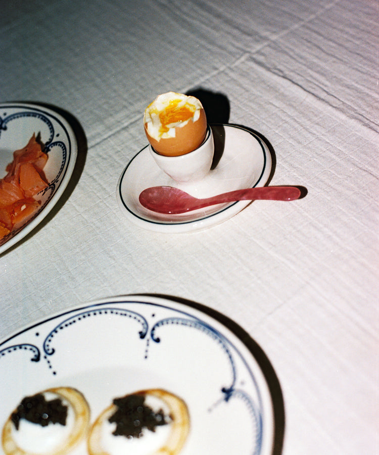 A soft-boiled egg in an egg cup with a pink Piccolo Spoon, beside plates of smoked salmon and blinis with cream and caviar on a white tablecloth. The dishwasher-safe Piccolo Spoon adds style and convenience to this elegant breakfast spread.