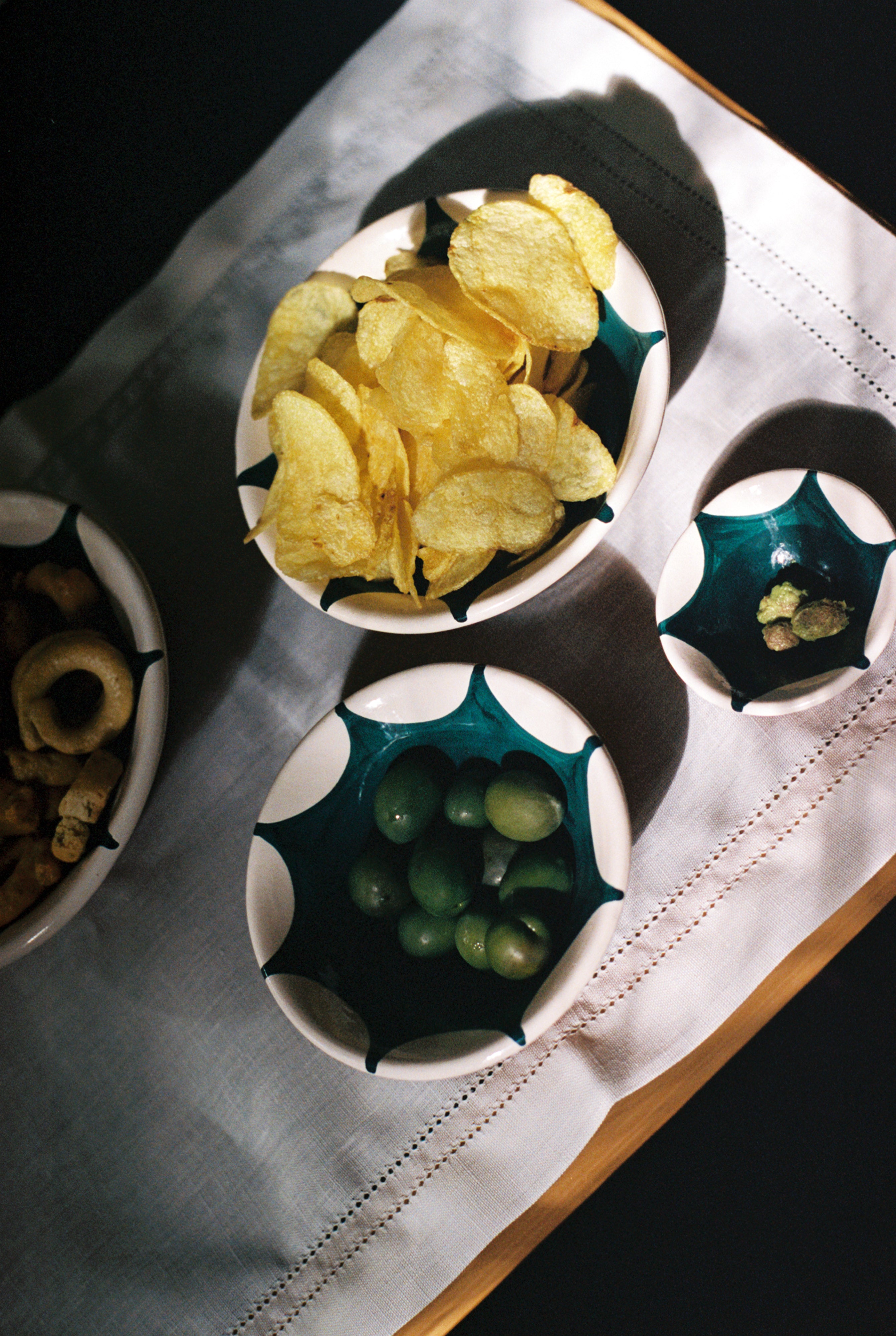 A bowl of potato chips, a bowl of green grapes, a bowl with a few nuts, and a bowl of mixed snacks are artfully arranged on a white cloth, each served in Stella Nesting Bowls that add charm and personality to your table setting.