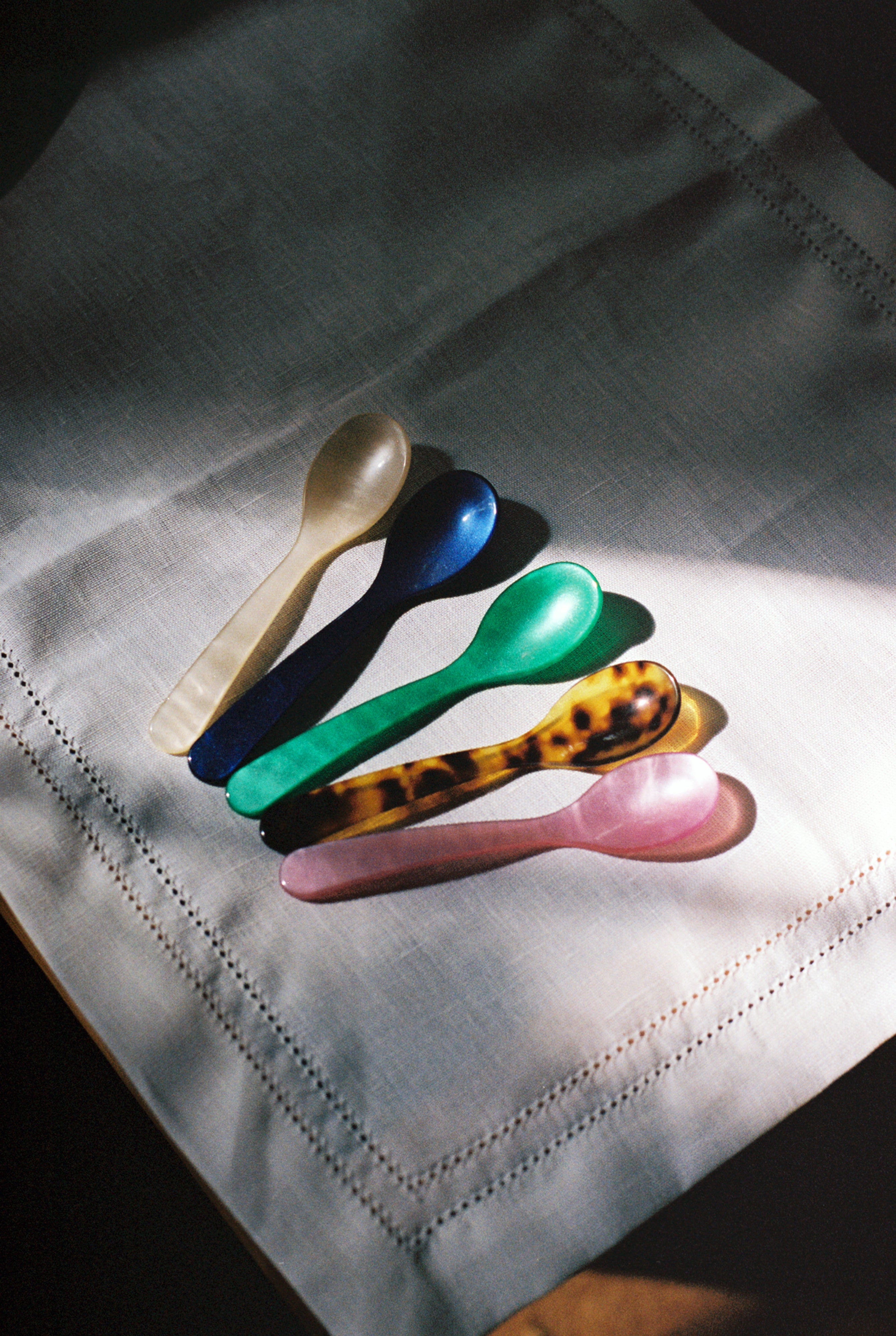 Five Piccolo Spoons—white, blue, green, tortoiseshell, and pink—display design craftsmanship as they lie side by side on a white linen napkin.