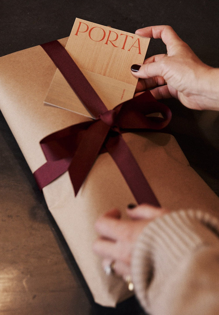 Person holding a gift box with a brown ribbon and 'Porta' card on a wooden surface