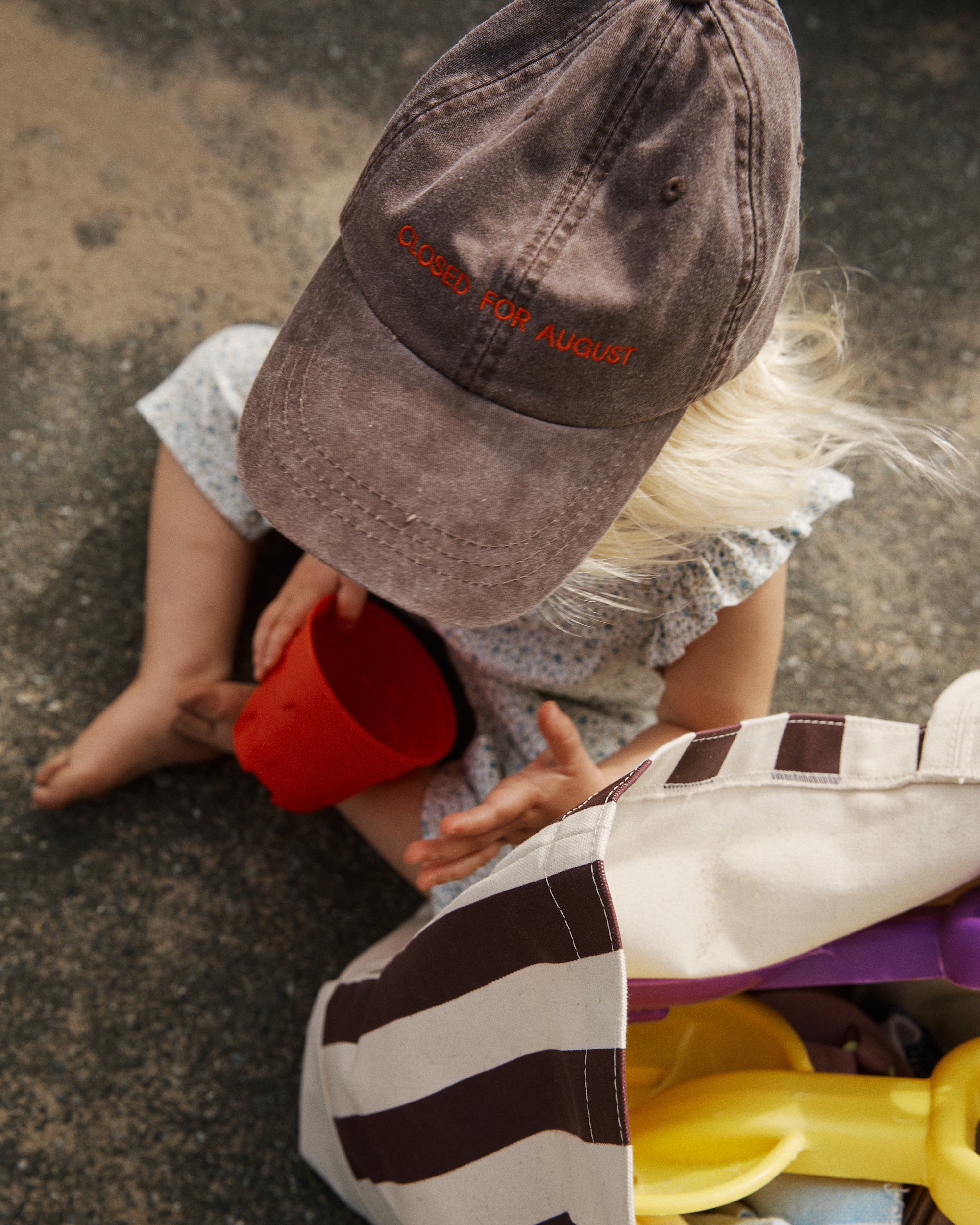 A child wearing the Closed for August Cap sits on the ground holding a red cup, with a striped bag of beach toys nearby.