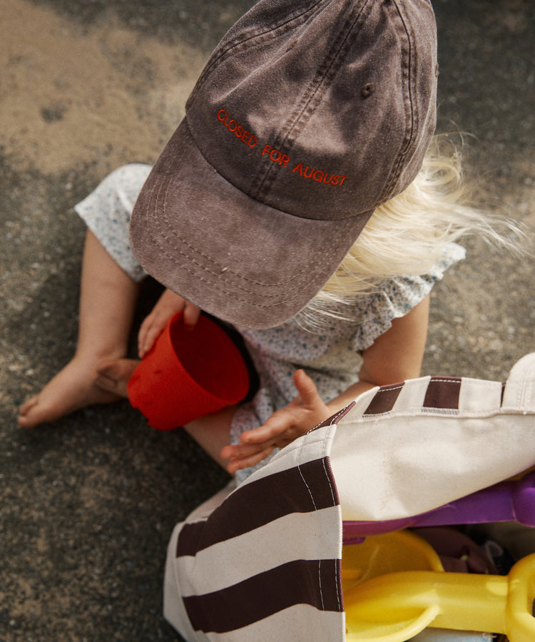 A child wearing the Closed for August Cap sits on the ground holding a red cup, with a striped bag of beach toys nearby.