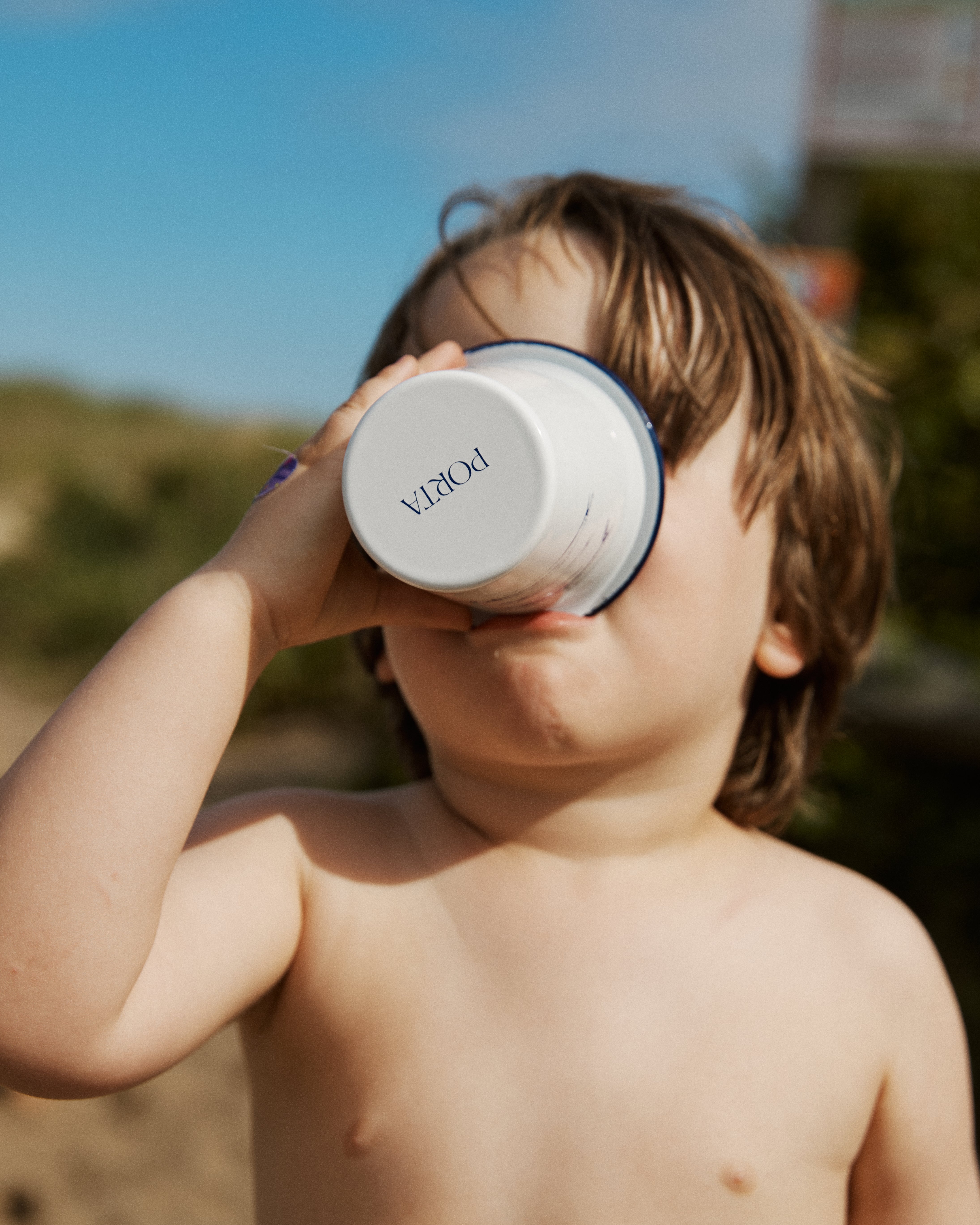 A young child with light brown hair drinks from the Il Pesce Enamel Cup outdoors, surrounded by greenery and a blue sky.