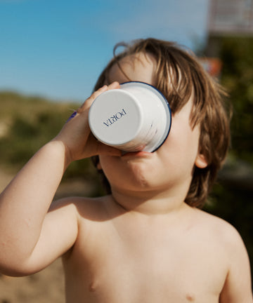 A young child with light brown hair drinks from the Il Pesce Enamel Cup outdoors, surrounded by greenery and a blue sky.