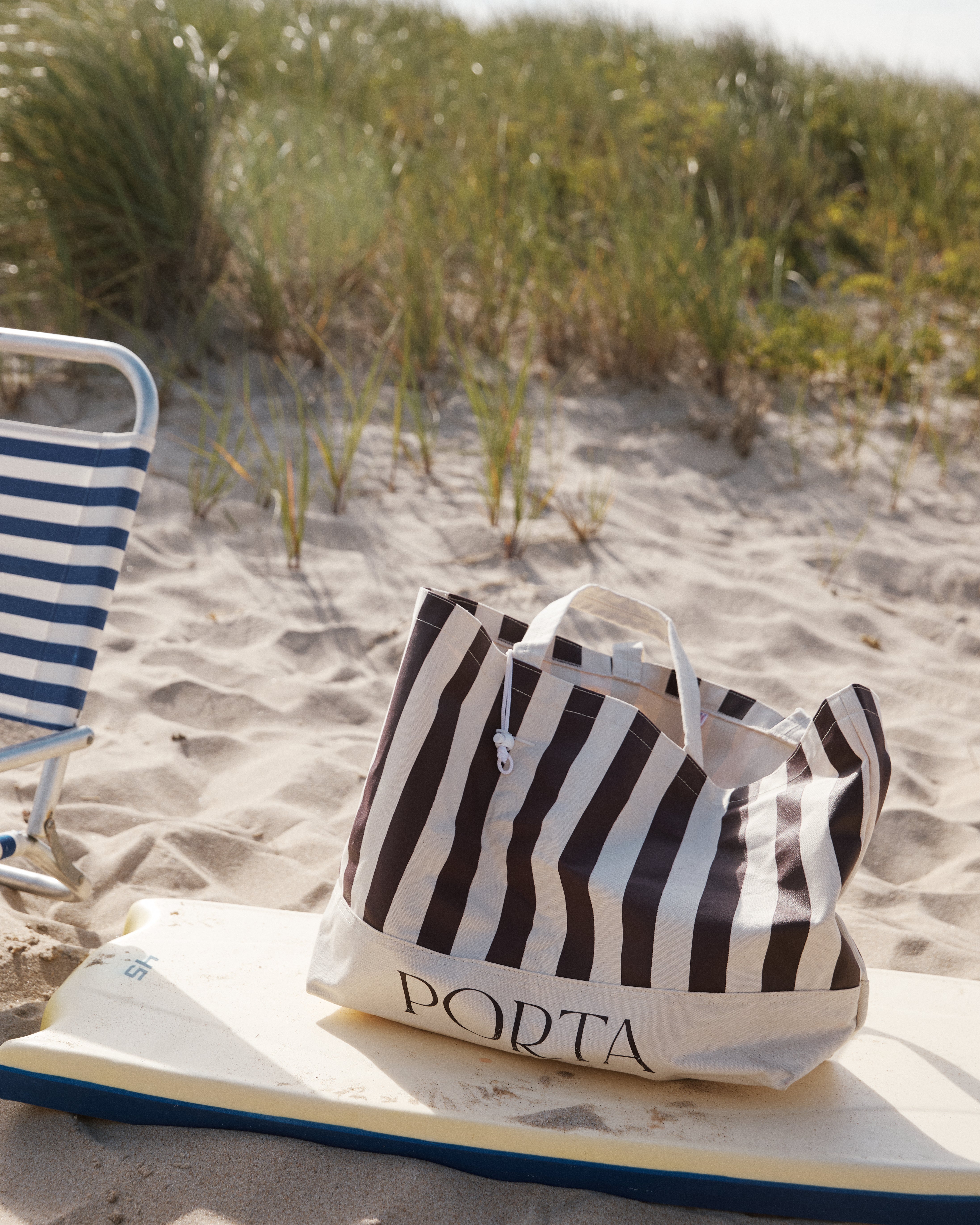 A PORTA Canvas Tote Bag with stripes sits on a surfboard next to a blue and white striped chair on a sandy beach with grass in the background.