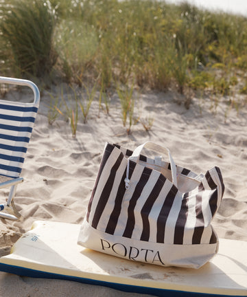 A PORTA Canvas Tote Bag with stripes sits on a surfboard next to a blue and white striped chair on a sandy beach with grass in the background.