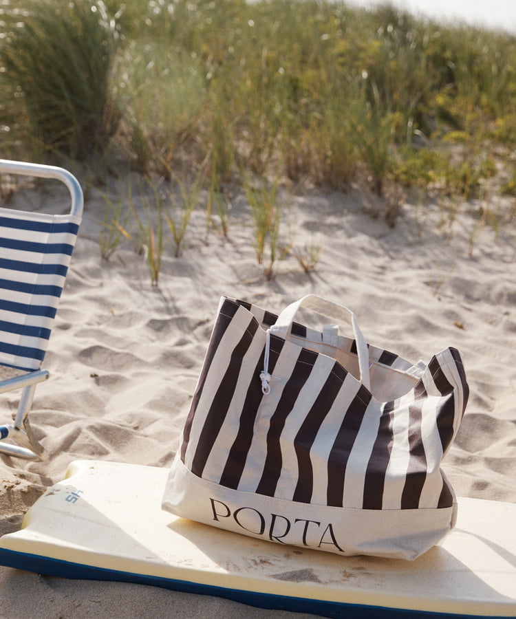 A PORTA Canvas Tote Bag with stripes sits on a surfboard next to a blue and white striped chair on a sandy beach with grass in the background.