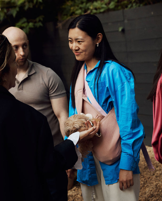 Woman holding a small dog in a sling while standing outdoors with other people.