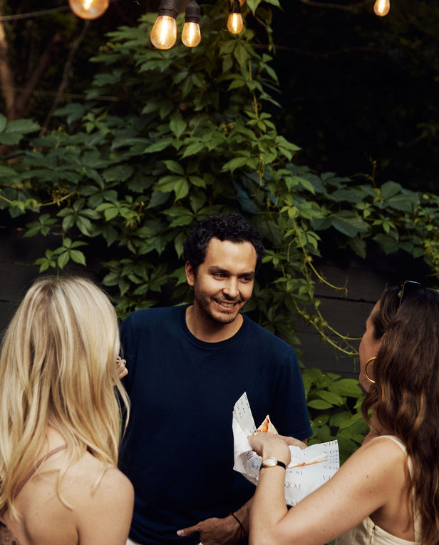 Three people standing outdoors with greenery and string lights in the background