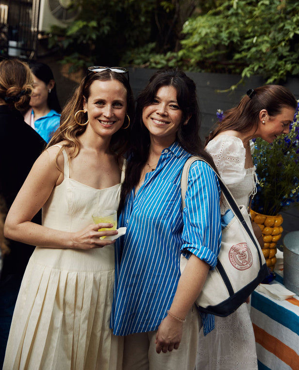 Two women standing together at an outdoor event, one holding a drink and the other with a backpack.