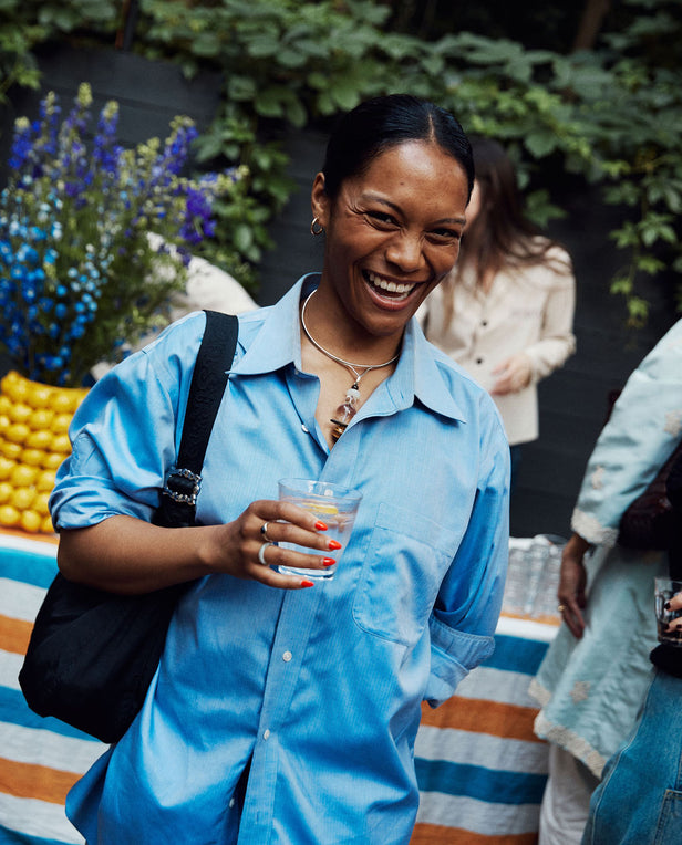 Woman in a blue shirt holding a drink, standing outdoors with greenery and colorful striped fabric in the background.
