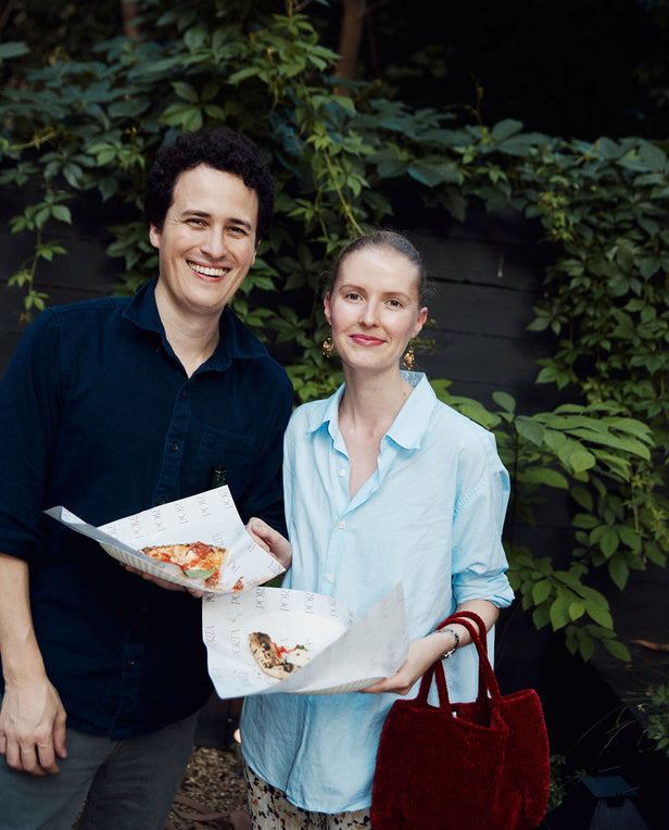 Two people holding pizza boxes outdoors with greenery in the background