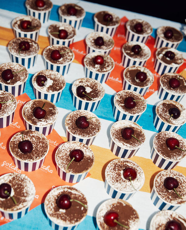 Small dessert cups with chocolate topping and a cherry on top, arranged on a colorful checkered tablecloth.