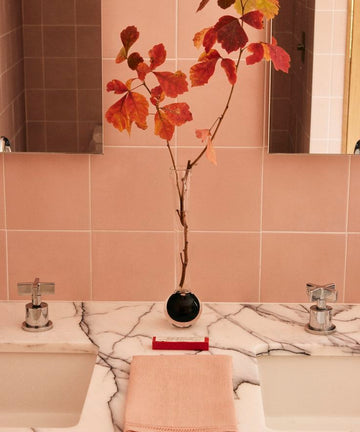 A marble countertop with two sinks features a folded pink towel, a red toothbrush, and the Pallino Bud Vase holding autumn leaves, all set against light pink tiled walls.