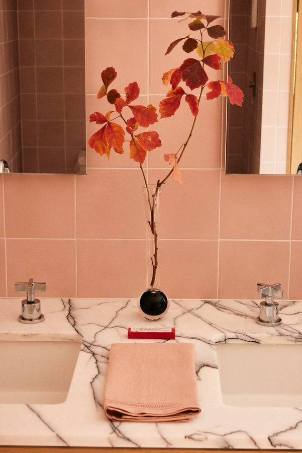A marble countertop with two sinks features a folded pink towel, a red toothbrush, and the Pallino Bud Vase holding autumn leaves, all set against light pink tiled walls.