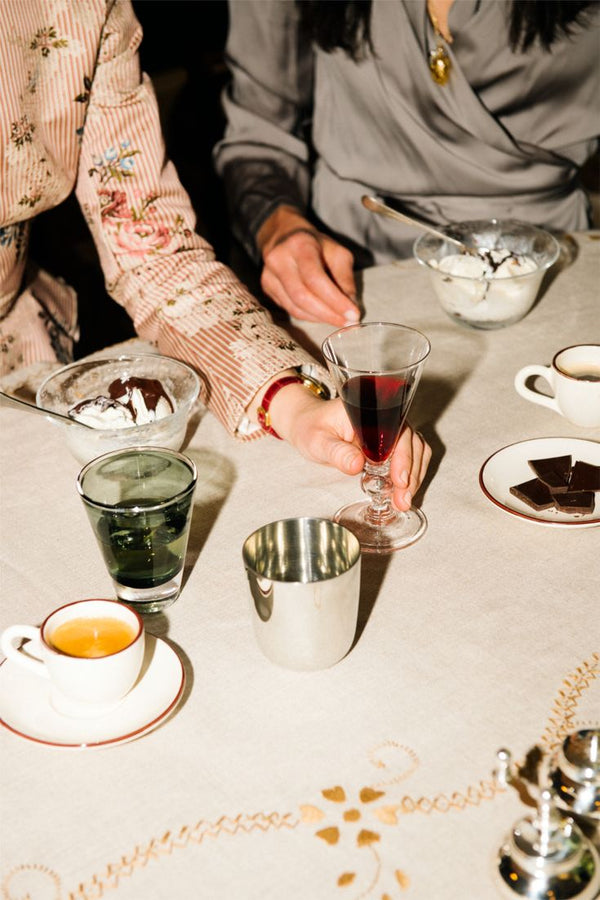 Two people sit at a table sipping wine from the Beata Stemmed Wine Glass, Set of 6, alongside water, coffee, bowls of ice cream, and chocolate on a cream-colored tablecloth.