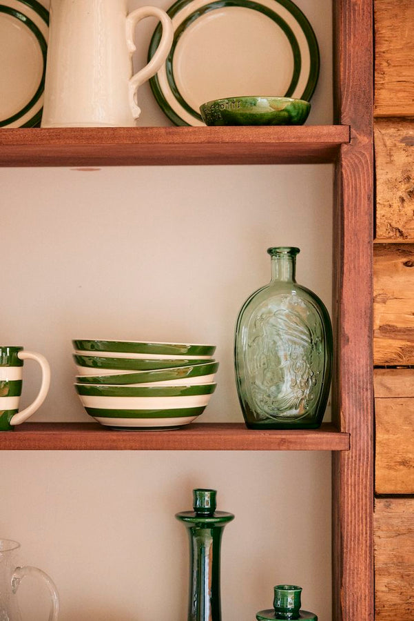 A wooden shelf showcases the Betty Cereal Bowl, green and white striped plates, a mug, a clear green glass bottle, and green glassware against a cream wall—an ideal setup for your breakfast collection.