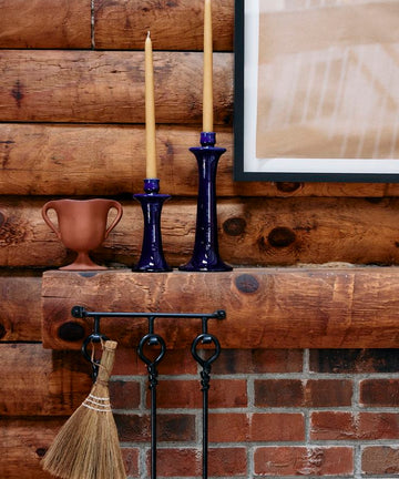 A wooden mantel displays two tall yellow candles in Maria Candleholders and a small brown vase, with fireplace tools and a broom hanging below against a brick and log wall.